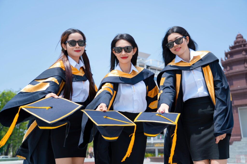 Three female graduates in gowns holding caps in celebration outdoors on a sunny day.